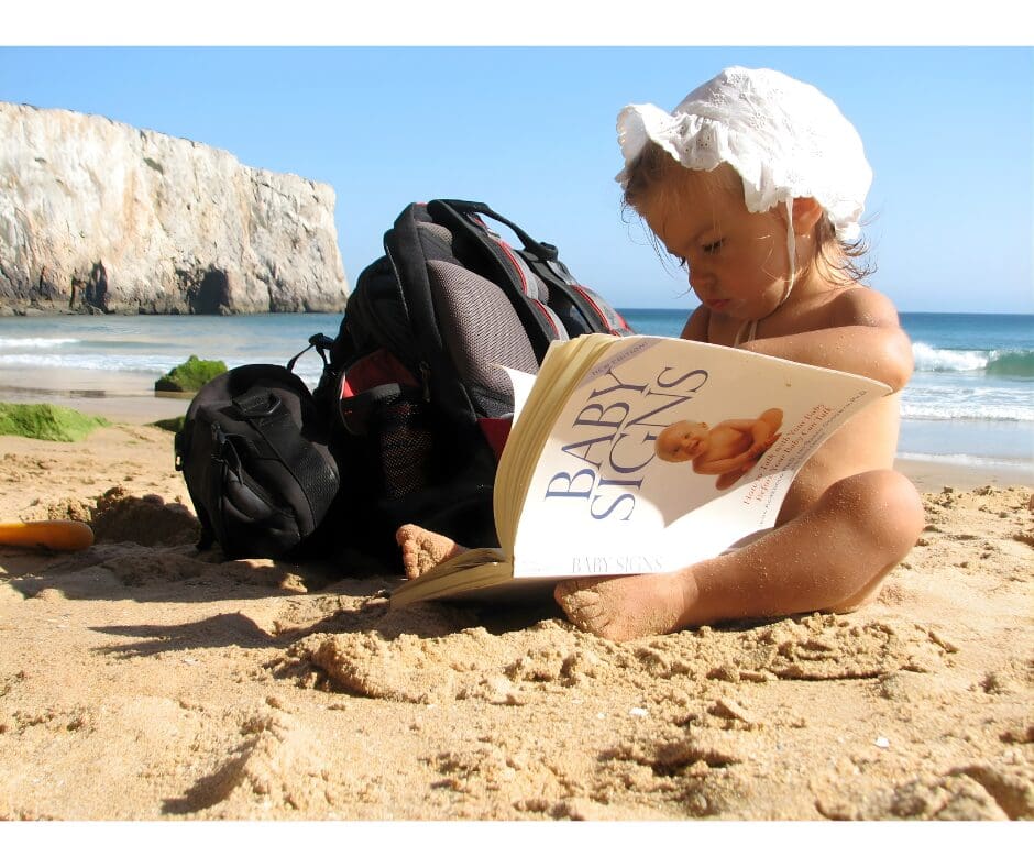 Baby sitting on the beach reading a board book, supporting early literacy development with Baby Signs®