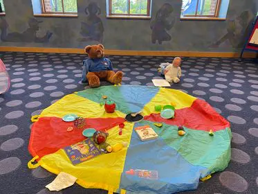 A teddy bear sits on a play mat in a room while being observed by a baby sign language teacher.