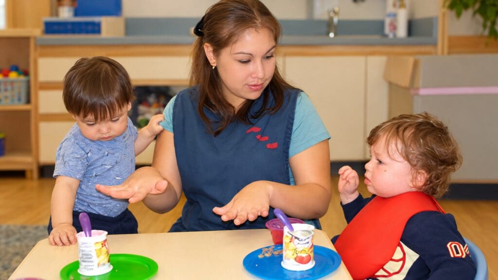Early childhood educator using hand gestures while interacting with two toddlers during mealtime.