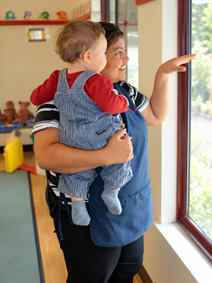 Early childhood educator holding a baby and pointing toward a window during a shared interaction.