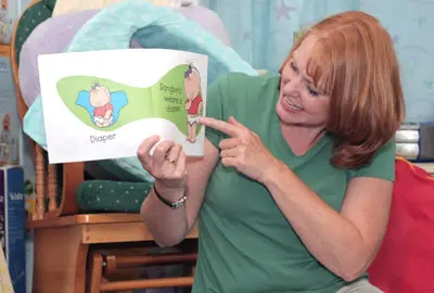 A woman sitting on a chair reading a book while teaching baby sign language.