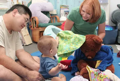 A group of people teaching a baby sign language while playing with a teddy bear.