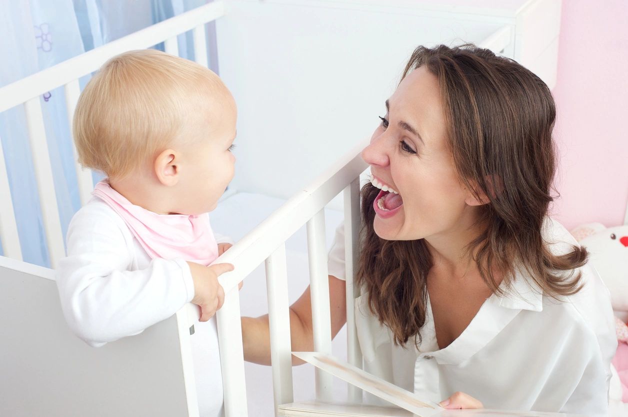 A joyful woman aiding language development by interacting with a baby in a crib in a brightly colored nursery.