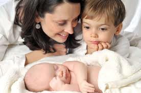 A mother and her young son smiling at a newborn baby, exhibiting early signs of communication development, sleeping under a white blanket.