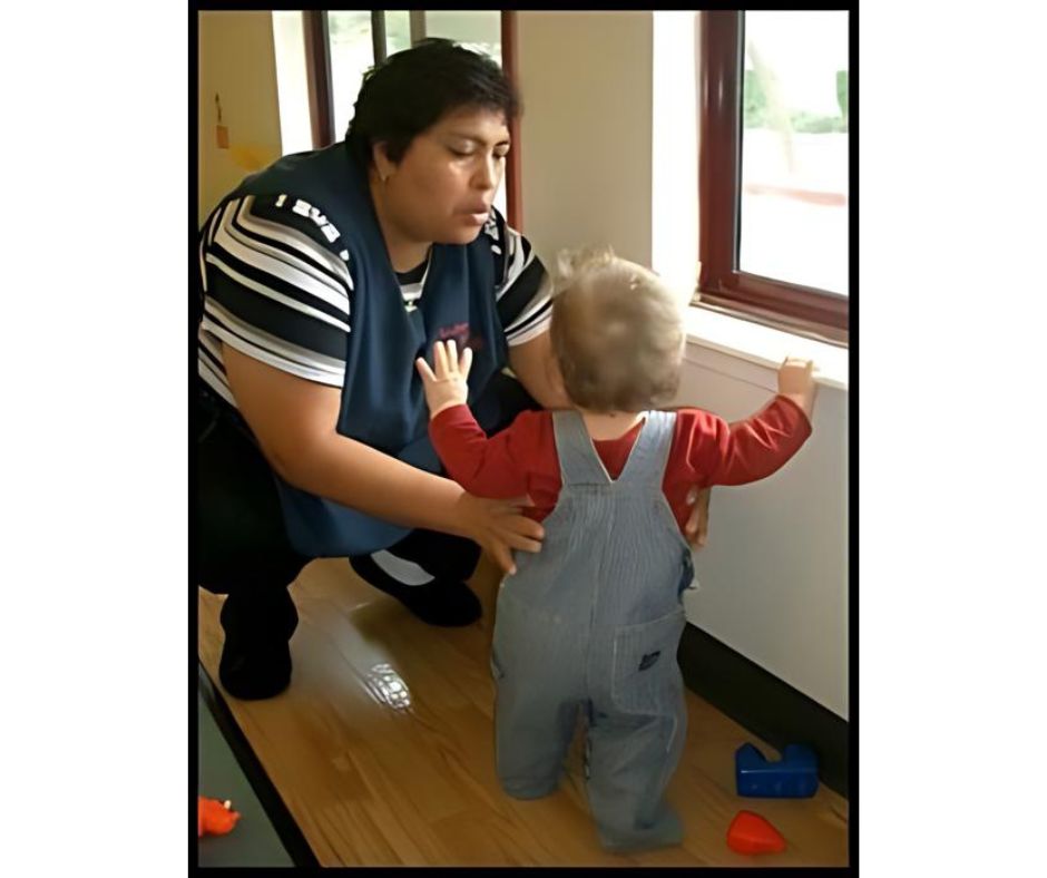 Woman helps toddler near window.