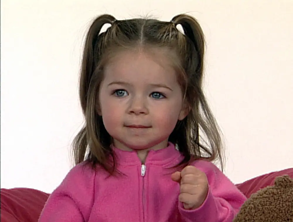 A toddler with pigtails wearing a pink jacket demonstrates the Baby Signs® sign for “milk” by opening and closing her fist.